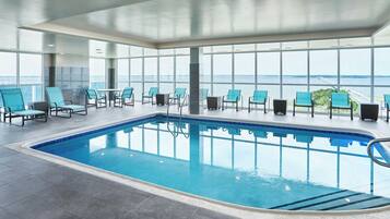 An indoor swimming pool with clear views of the sea and a bridge, surrounded by blue chairs and a tiled floor.