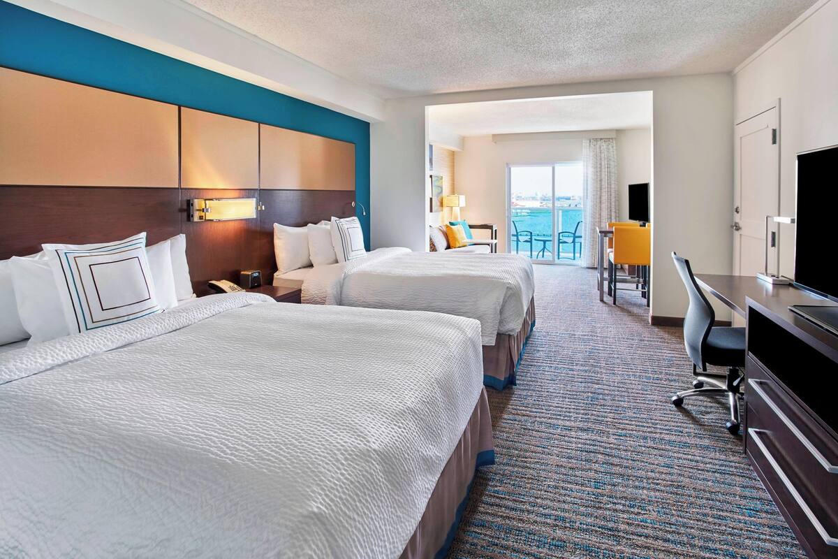 A hotel room with two beds, a desk, and a TV. There is a balcony with a view of the city.