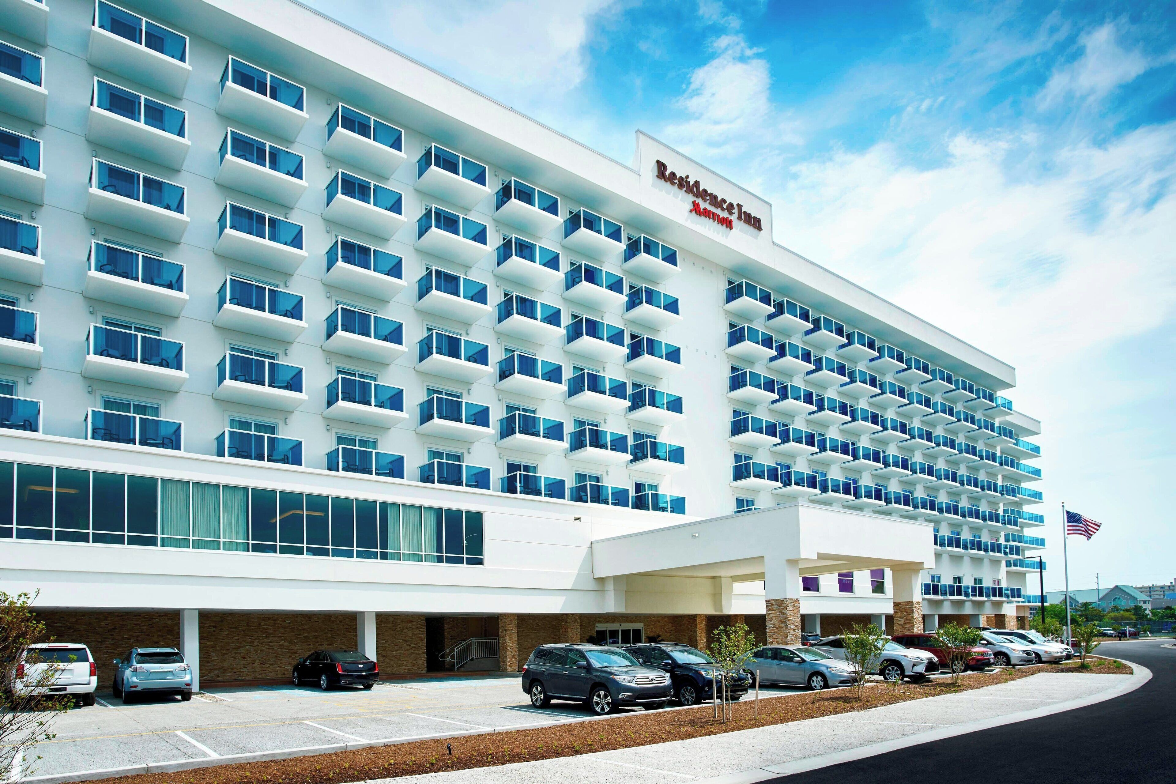 A modern hotel with a white facade, multiple balconies, and a parking lot.