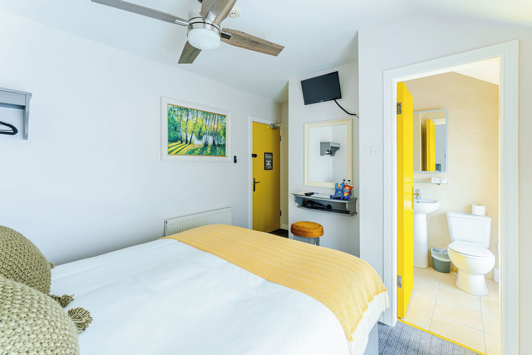 A hotel room with a bed, a ceiling fan, a painting, a television, and a bathroom visible through an open door.