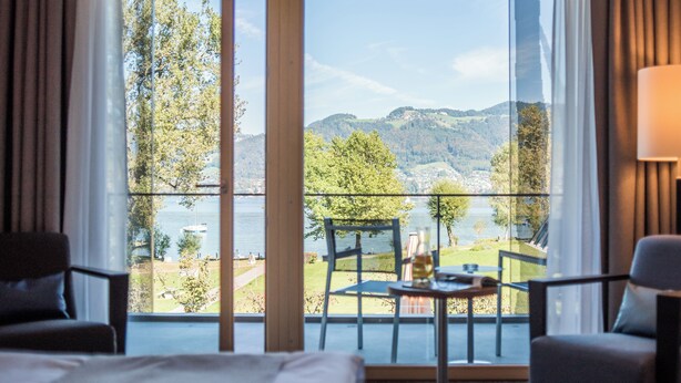 A hotel room with a large window offering a view of a lake and mountains. There is a bed, two chairs, and a small table with a bottle and glasses.