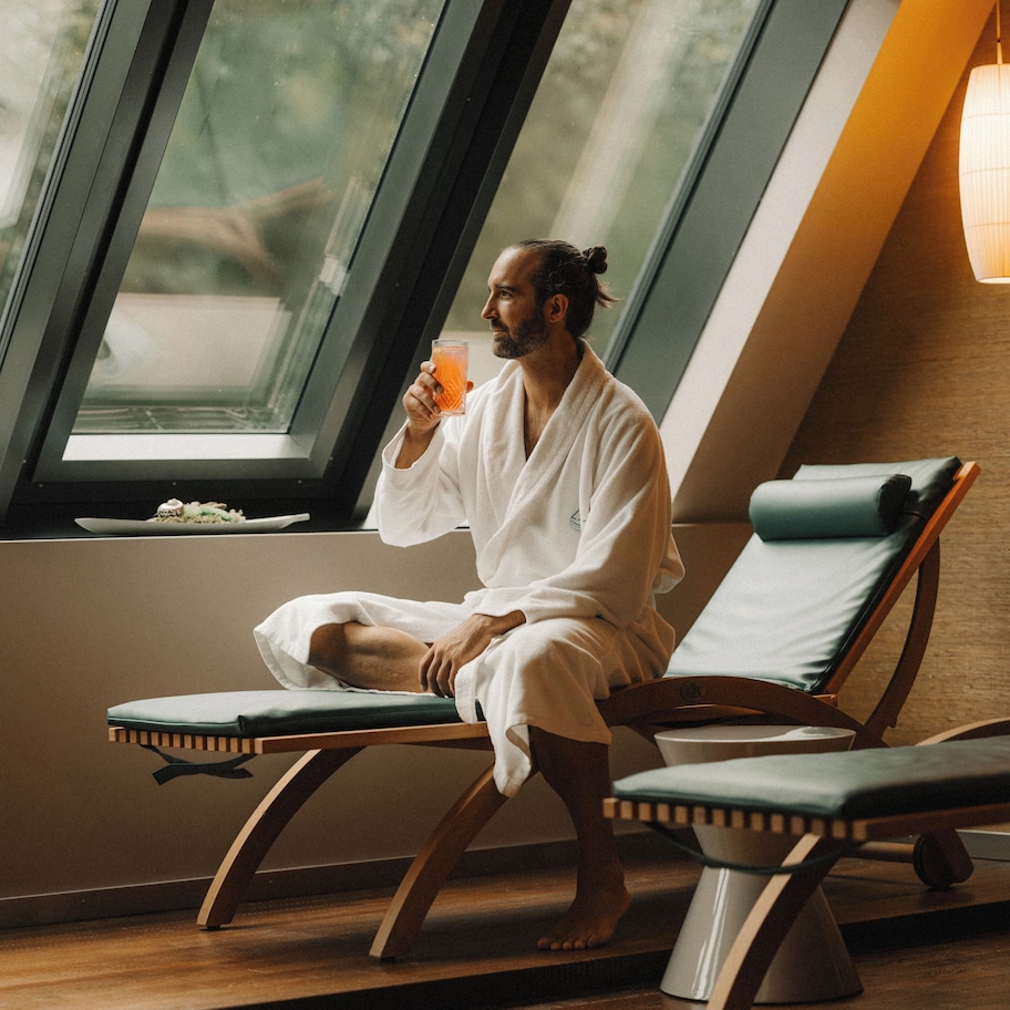 A person in a white bathrobe sitting on a wooden lounge chair by a window with a view of greenery outside.