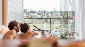 A couple in a bathtub with a view of a residential area through a large window.