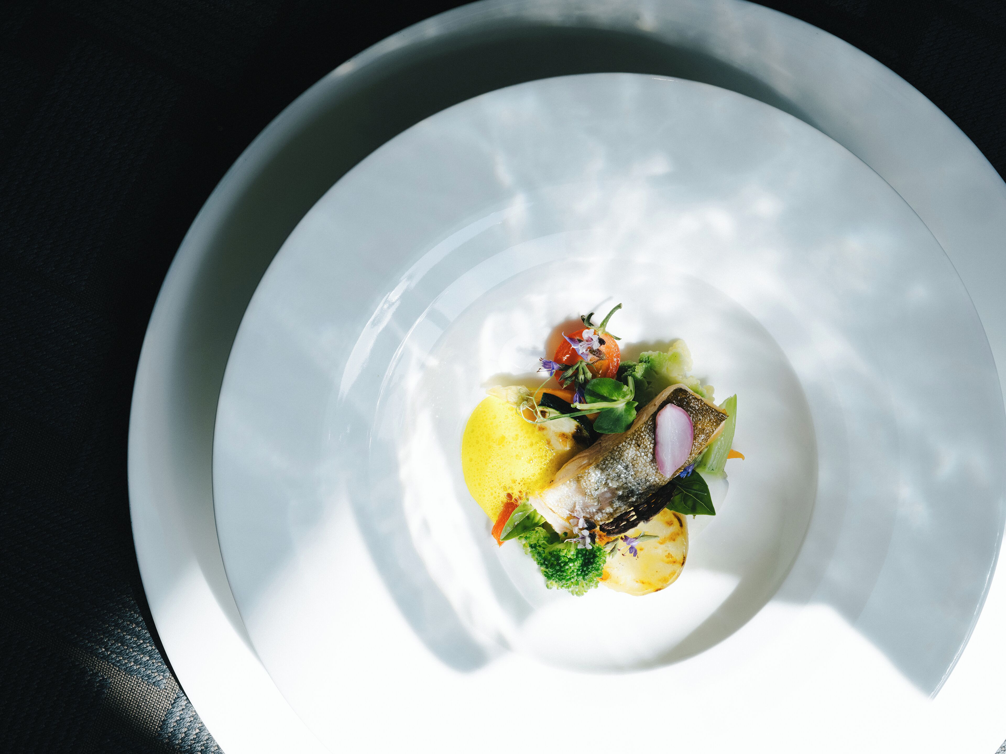 A white plate with a fish, vegetables, and a yellow item, possibly a potato, on a dark surface.