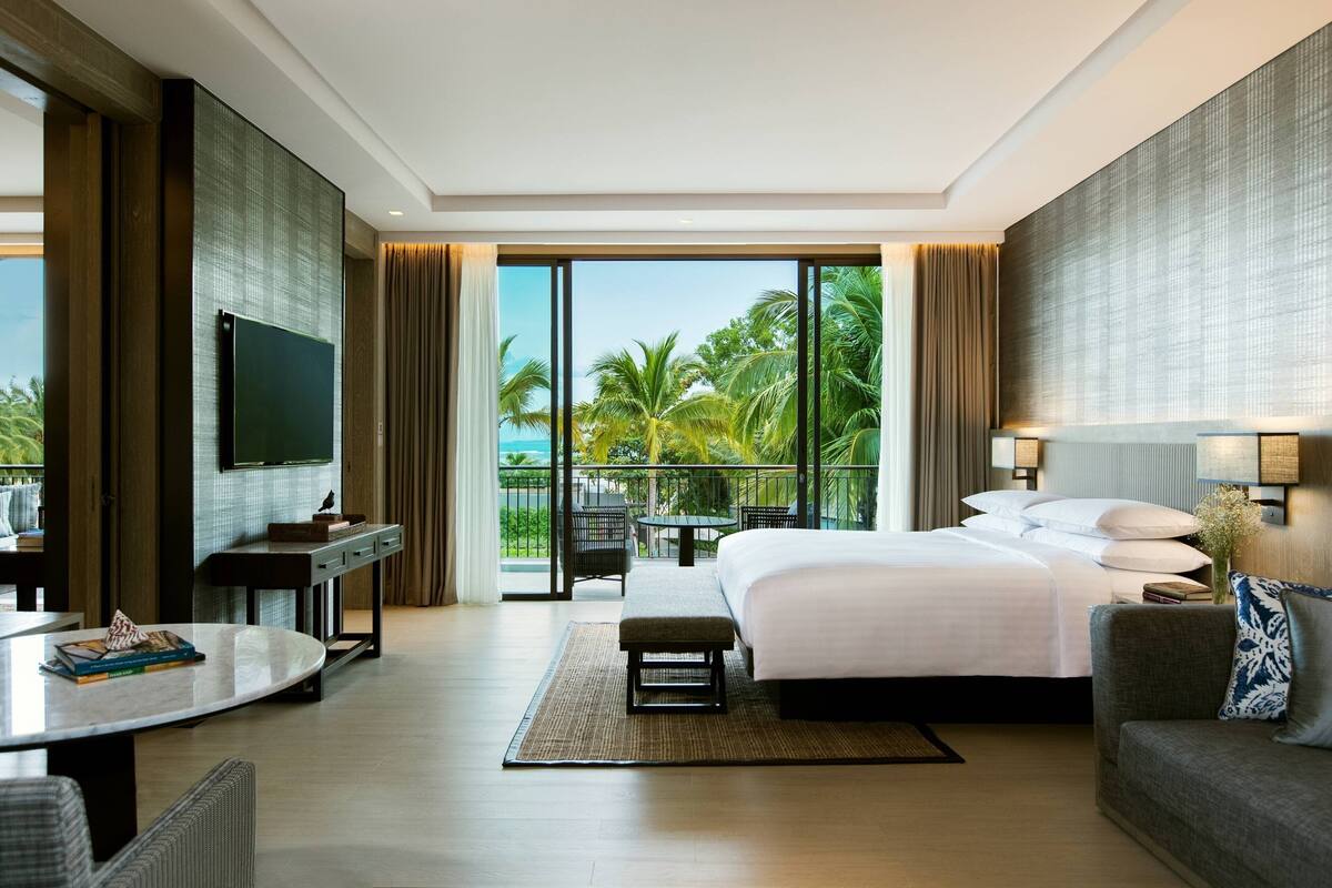 A hotel room with a large bed, a desk with a flat-screen TV, a balcony with a view of palm trees, and a dining area with a table and chairs.