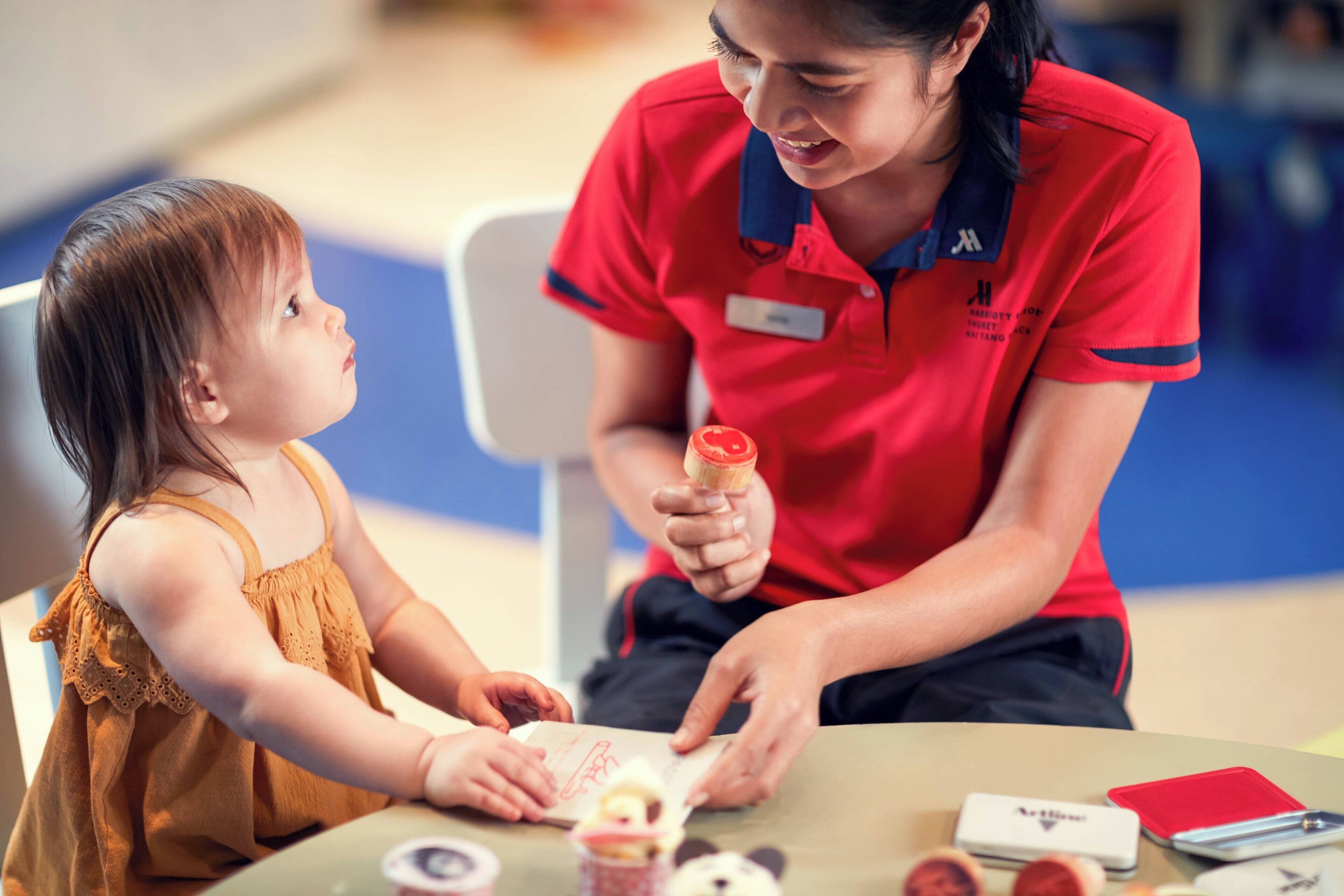 Una mujer con uniforme rojo sentada a una mesa con un niño, sosteniendo un objeto pequeño.
