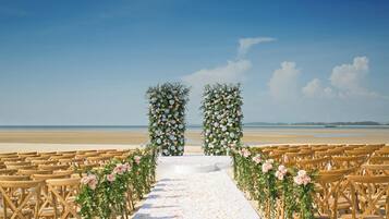 A beach wedding setup with a white aisle, floral arches, and wooden chairs.