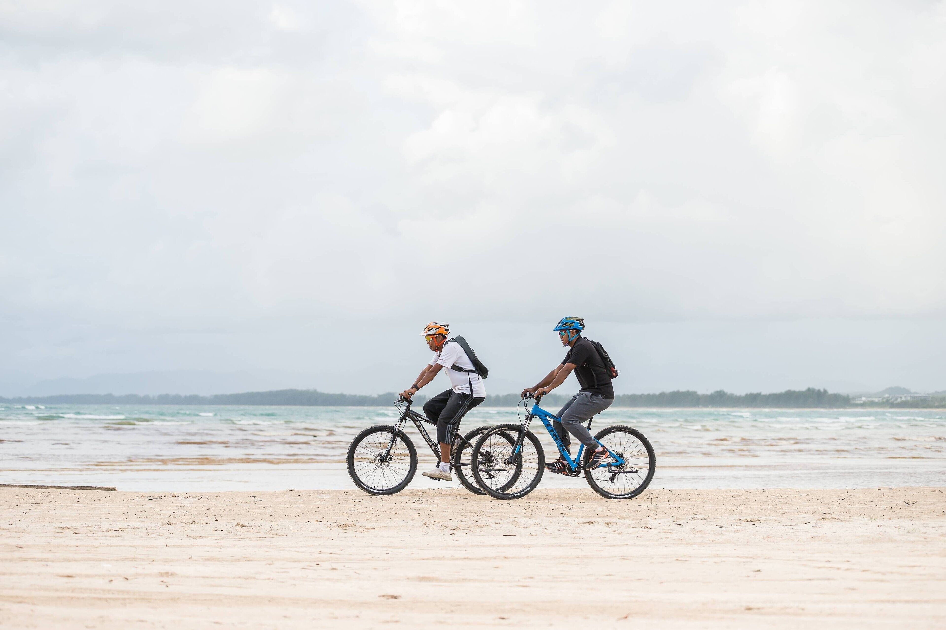 Two cyclists on a beach with a view of the ocean and distant hills.
