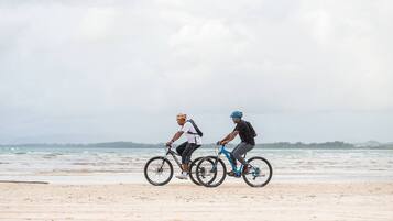 Two cyclists on a beach with a view of the ocean and distant hills.