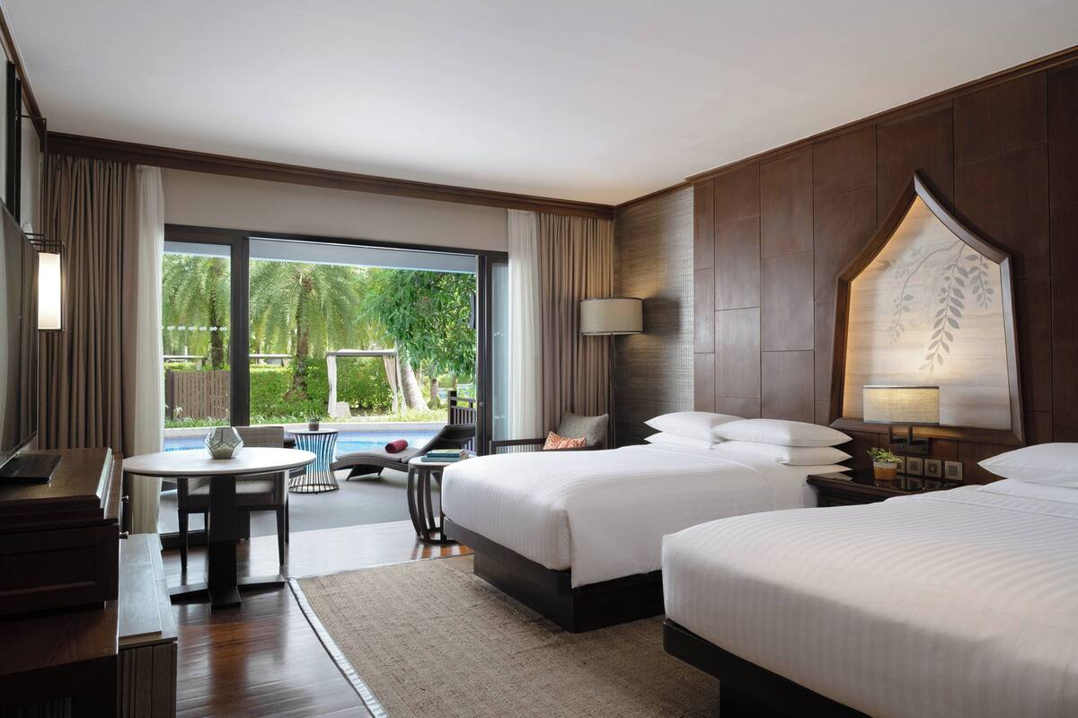 A hotel room with two beds, a desk, a chair, and a view of a swimming pool and greenery.