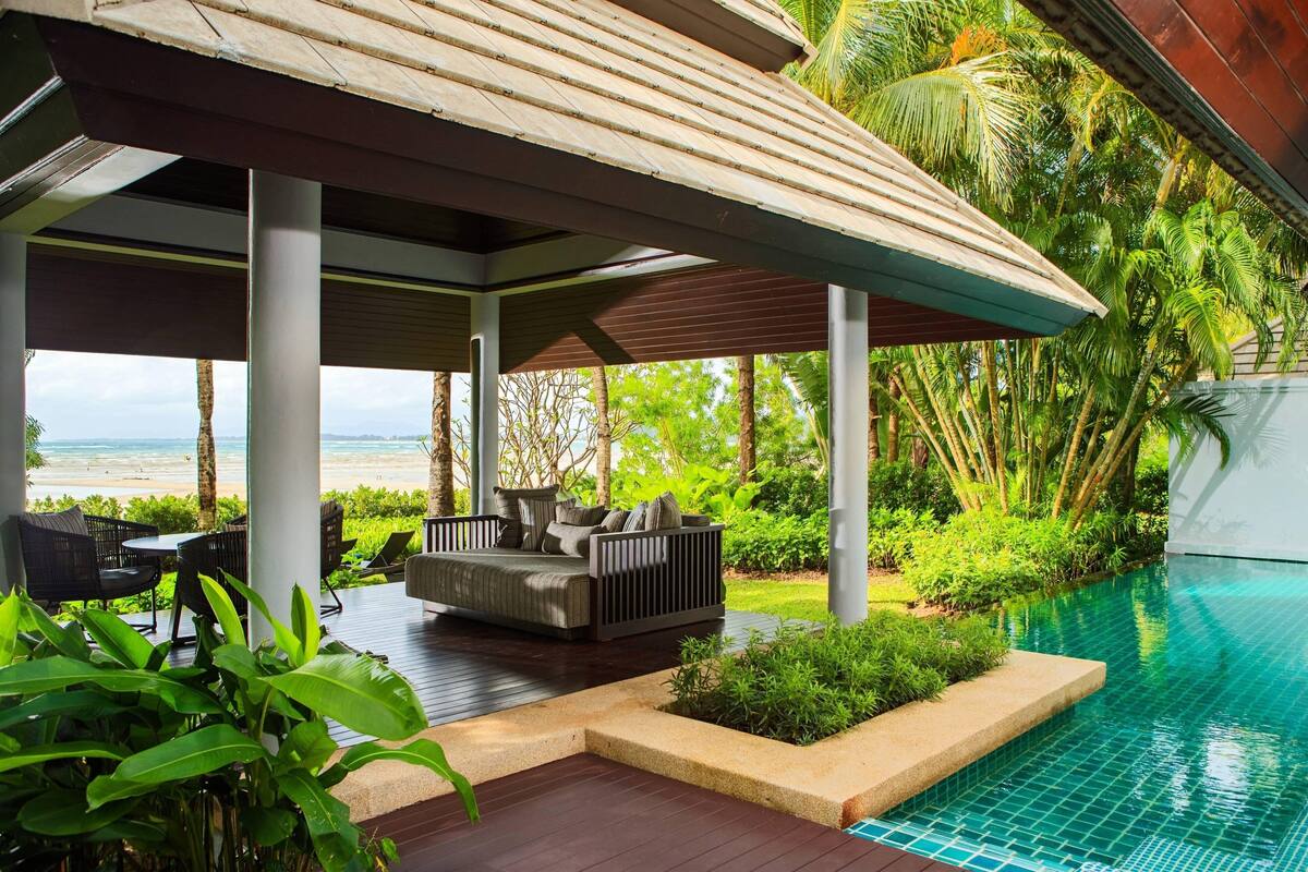 A covered outdoor seating area with a pool, surrounded by lush greenery and a view of the beach.