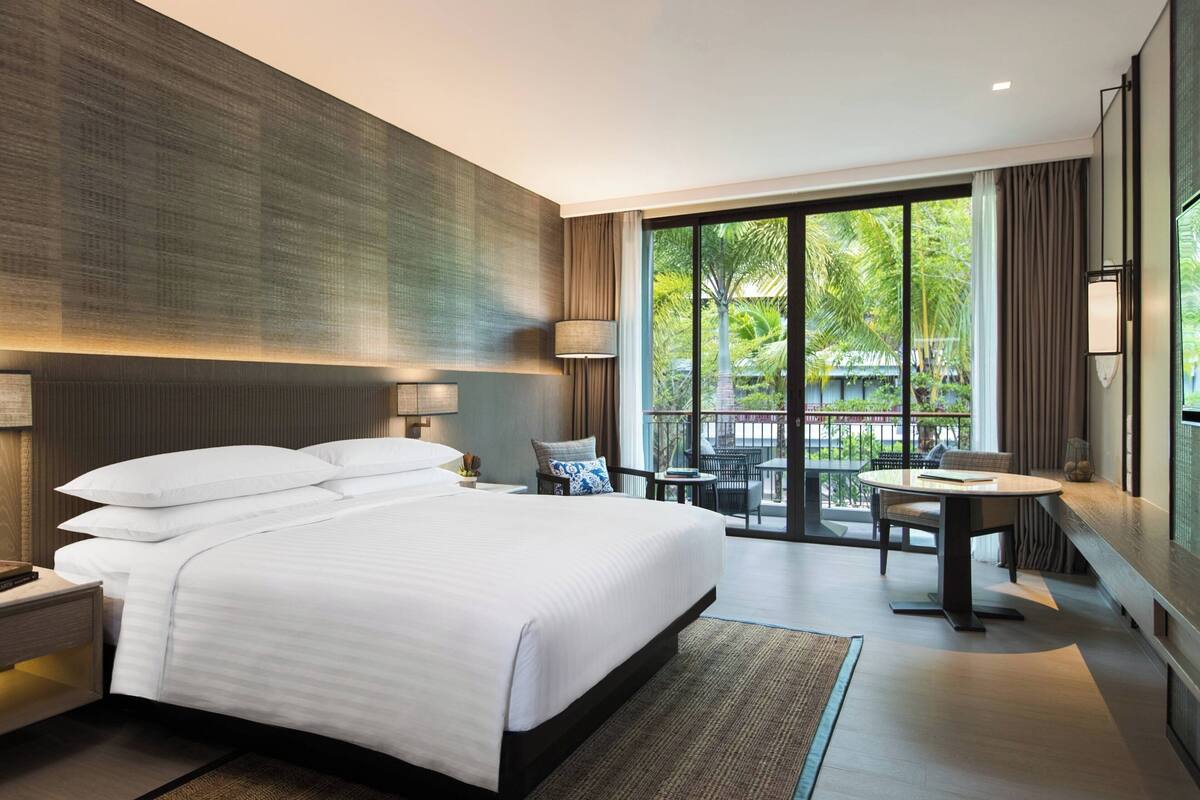 A modern hotel room with a large bed, a desk, and a balcony with a view of greenery.