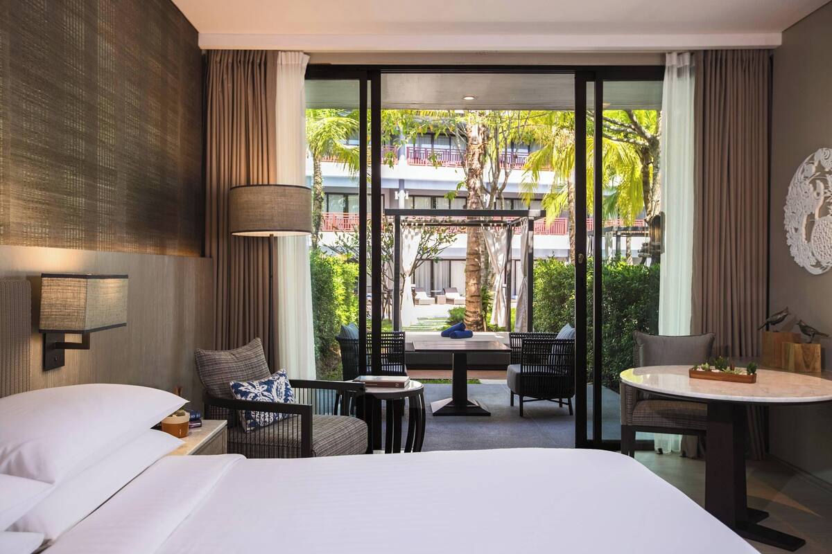 A hotel room with a bed, two chairs, a small table, and a view of a courtyard with plants and a building.