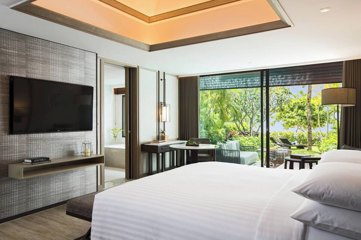 A modern hotel room with a large bed, a flat-screen TV mounted on the wall, a small desk with a chair, and a view of lush greenery outside.