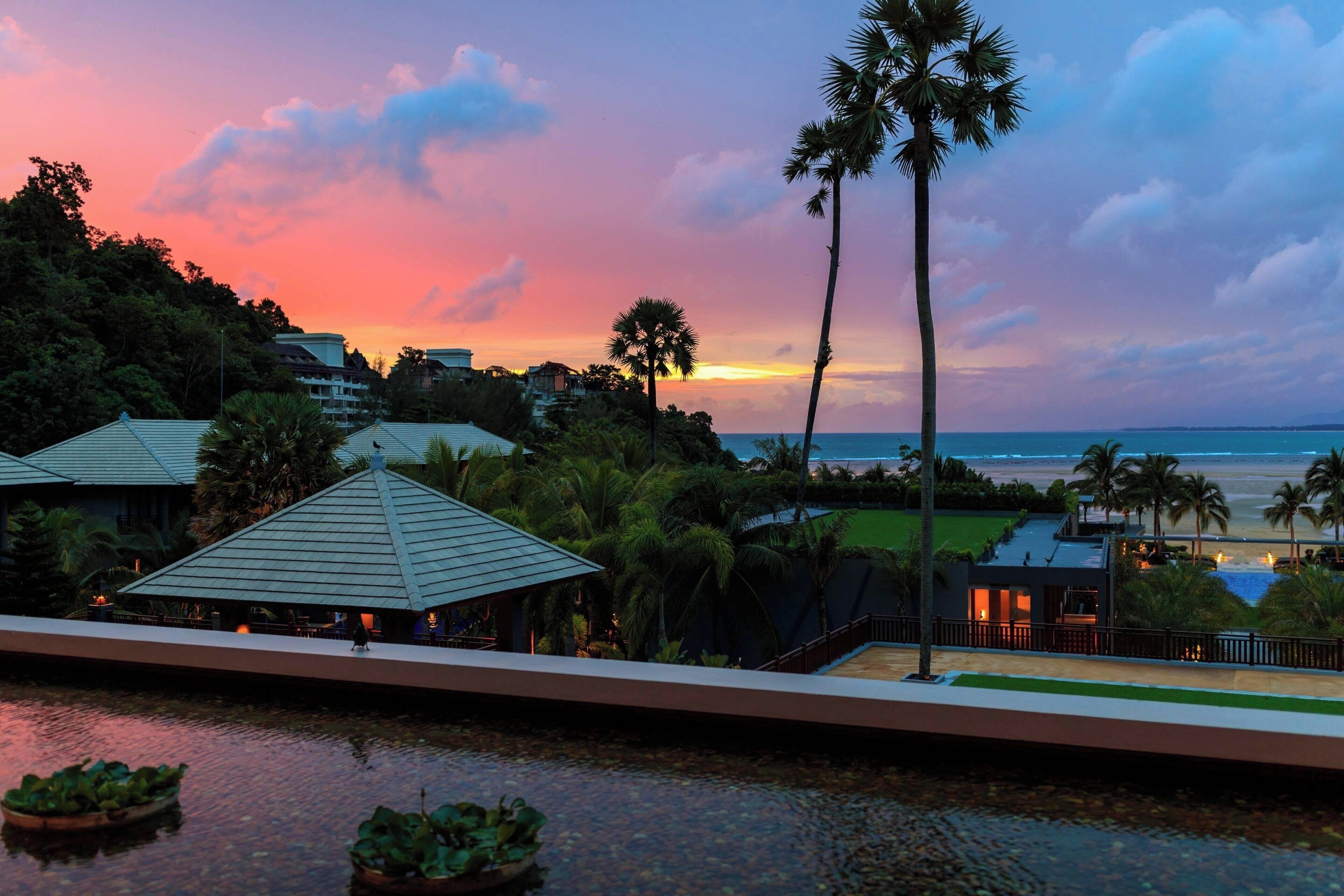 A resort with a view of the ocean at sunset, palm trees, and a pool area.