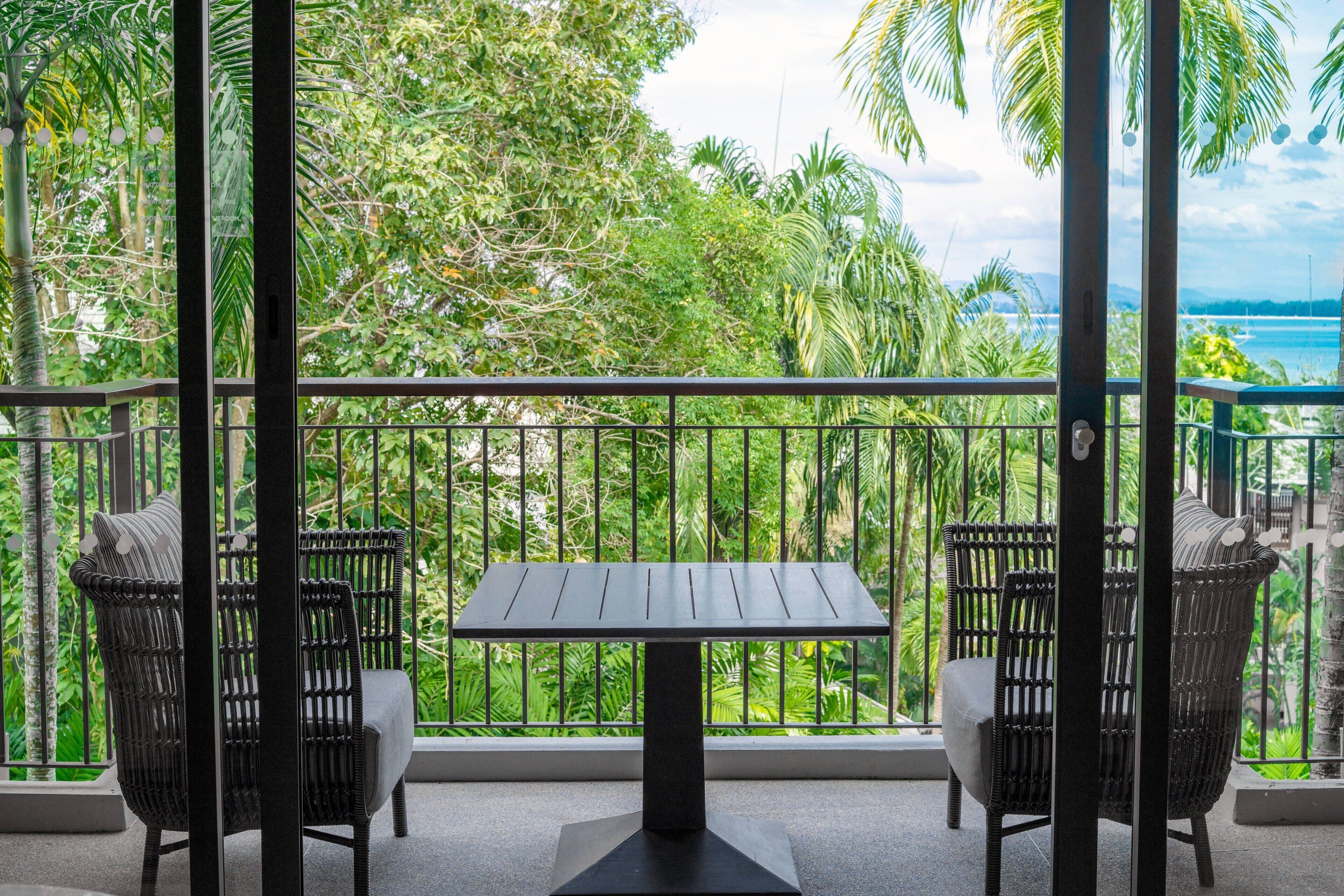 Un balcón con dos sillas de mimbre y una mesa, con vistas a un jardín exuberante y el mar.