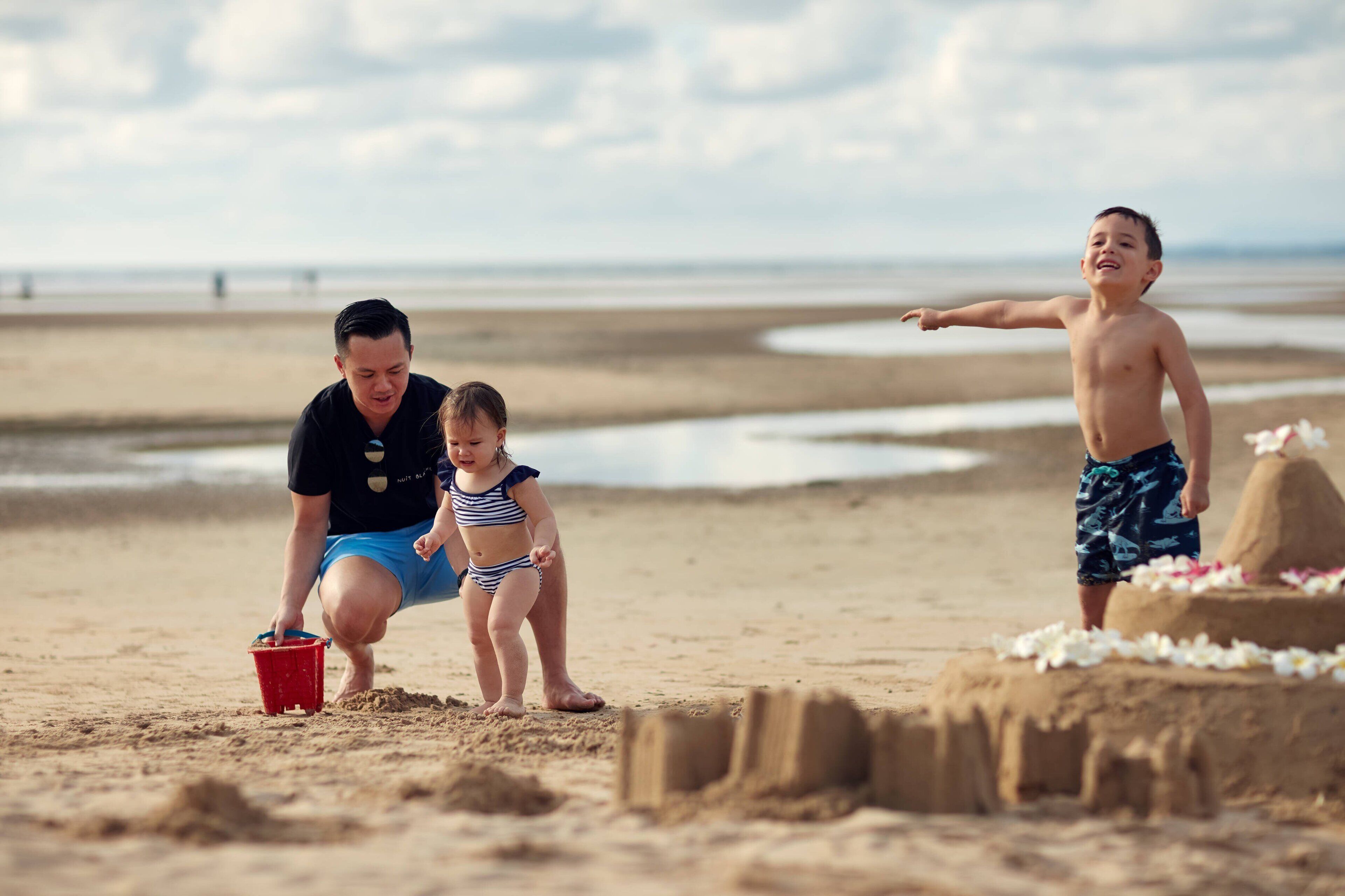 Escena de playa con un castillo de arena, un cubo rojo y dos niños jugando.