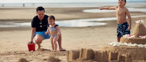 Escena de playa con un castillo de arena, un cubo rojo y dos niños jugando.