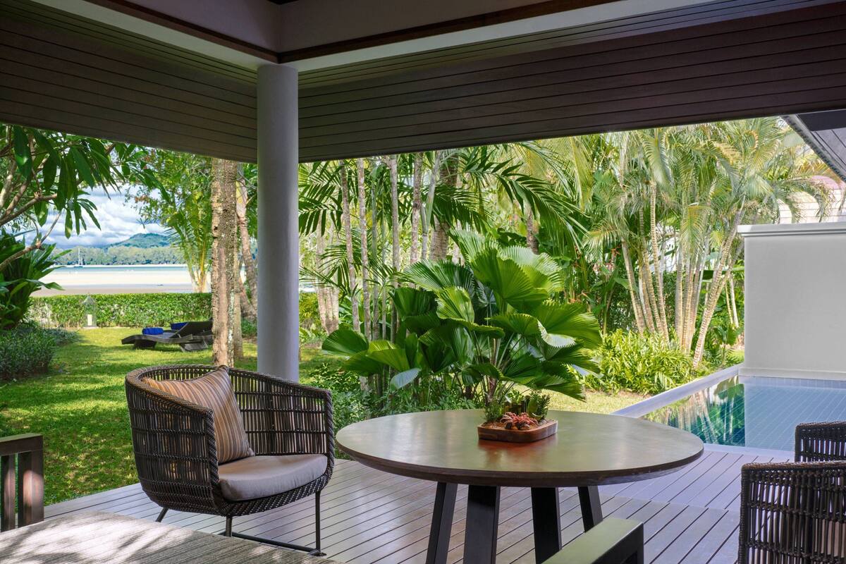 A covered outdoor area with a round table and wicker chairs, overlooking a pool and lush greenery.