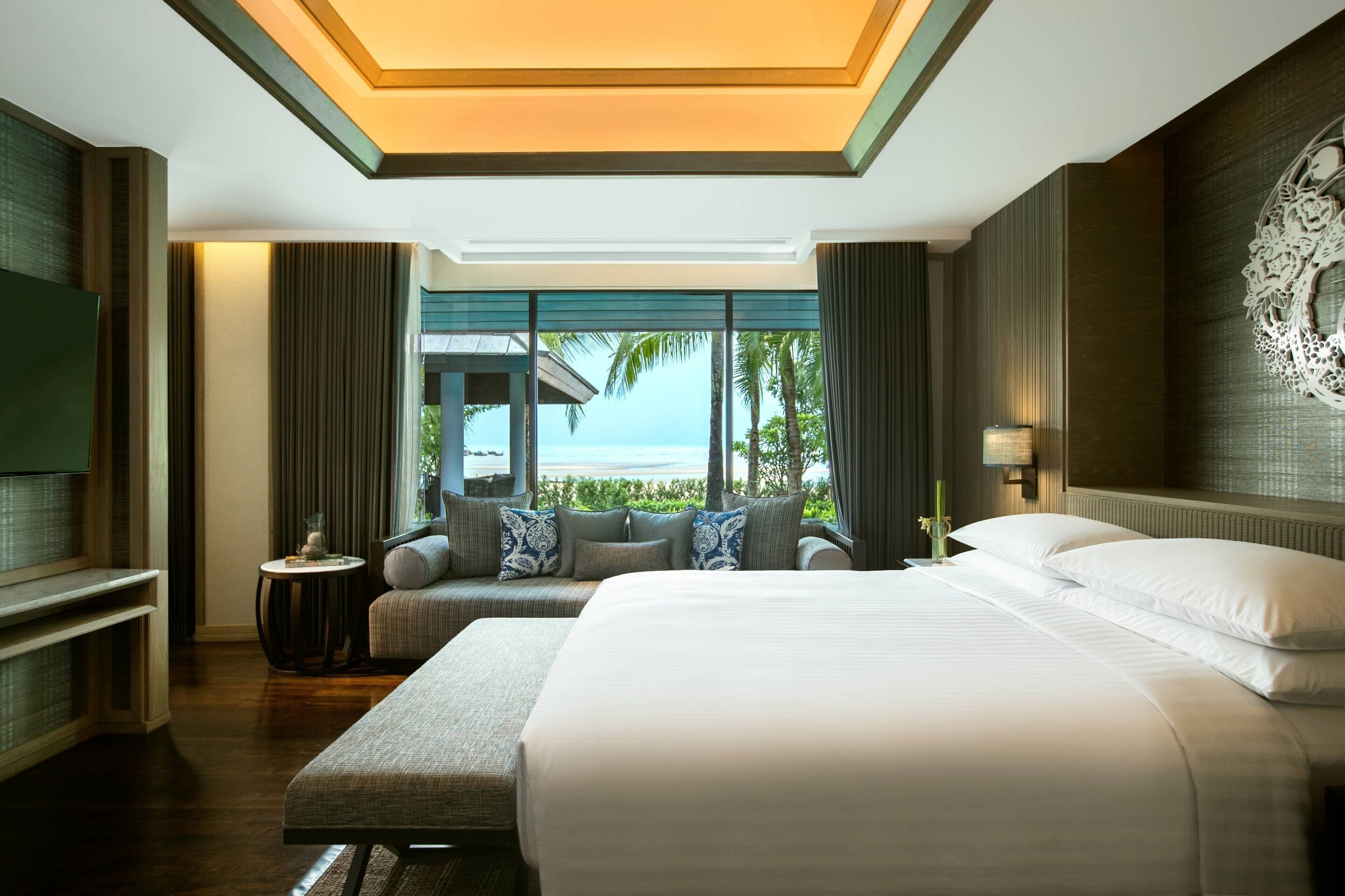 A hotel room with a large bed, a sofa, a small table, and a TV. The room has a view of the ocean.