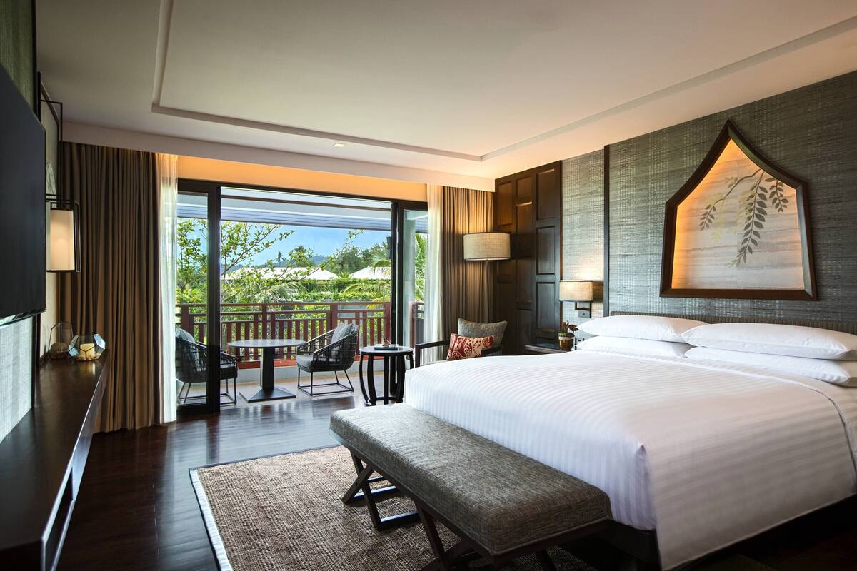 A modern hotel room with a large bed, a bench, a desk, and a balcony with outdoor furniture.