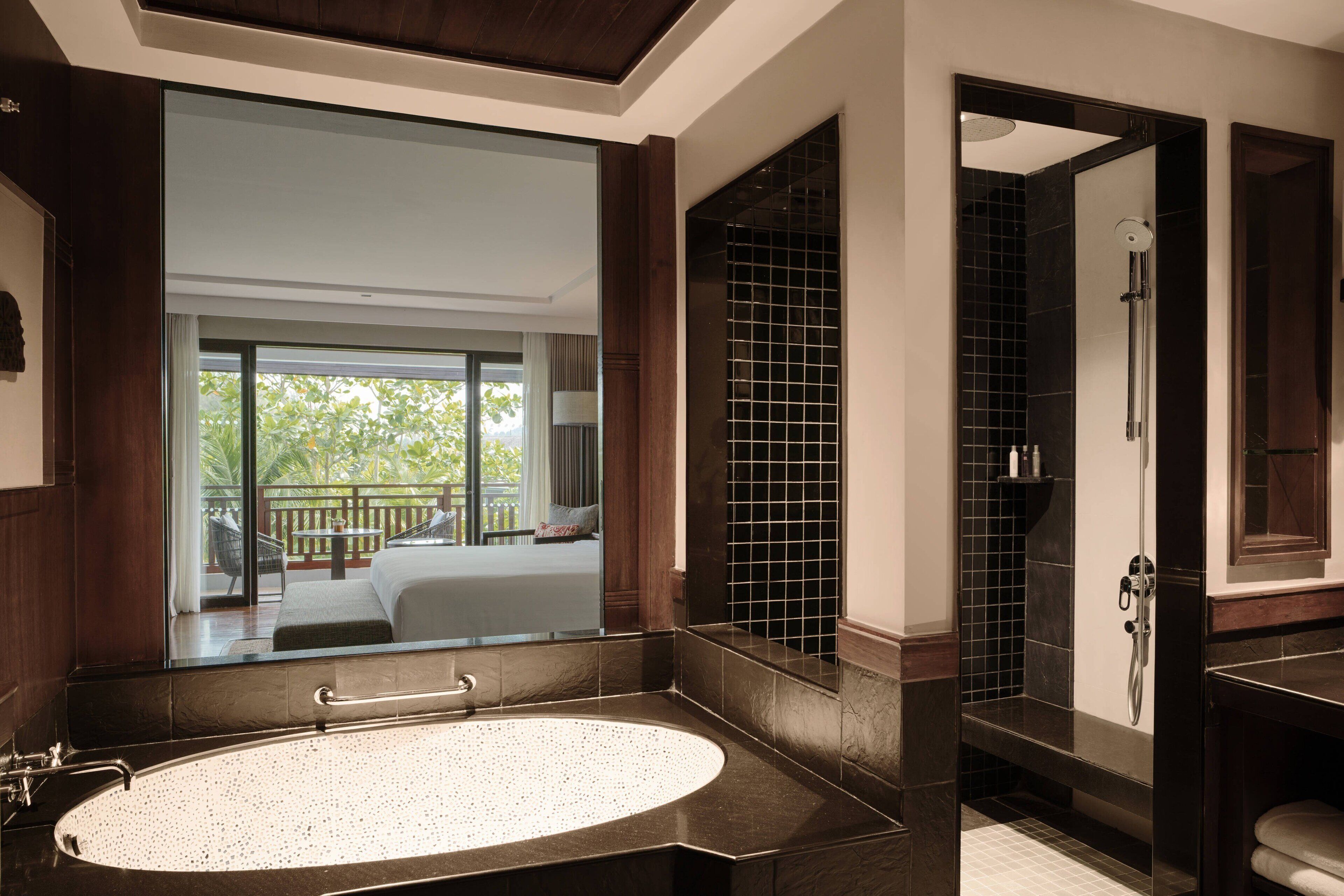 A modern bathroom with a large oval sink, a glass-enclosed shower, and a large mirror reflecting a balcony with outdoor furniture.