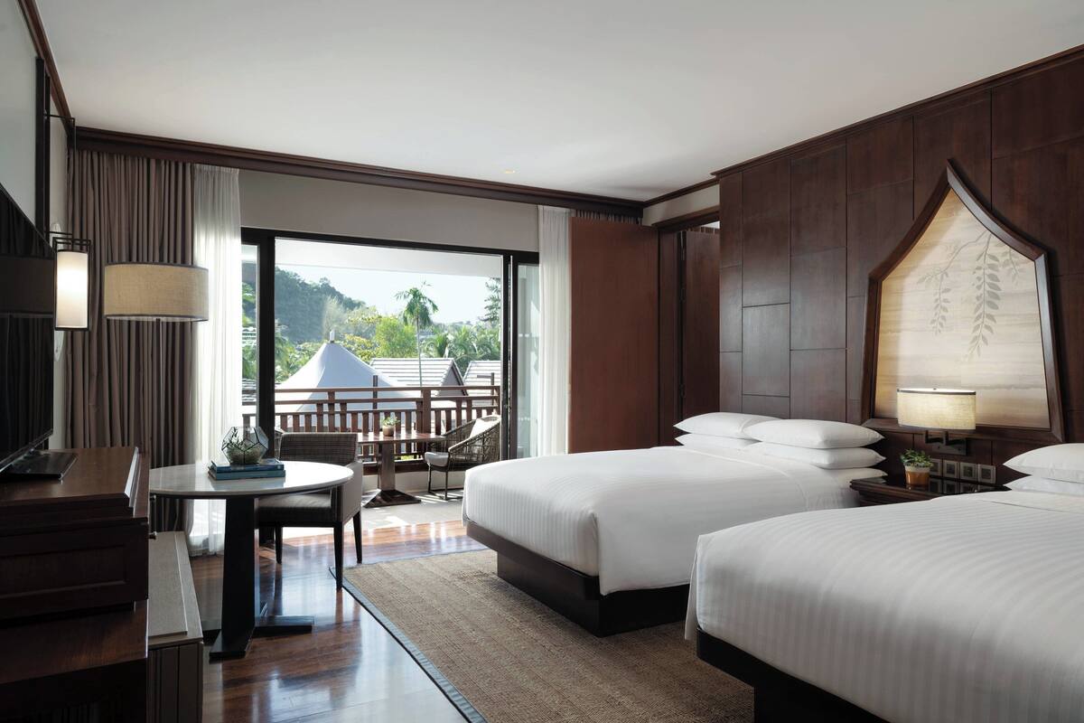 A hotel room with two beds, a desk, a chair, and a balcony with a view of greenery.