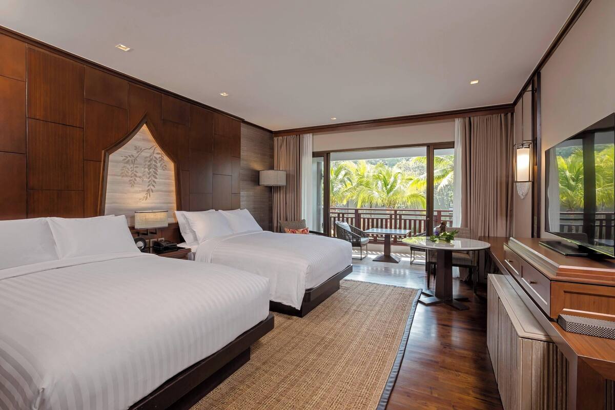 A hotel room with two beds, a dining area, a TV, and a balcony with a view of palm trees.