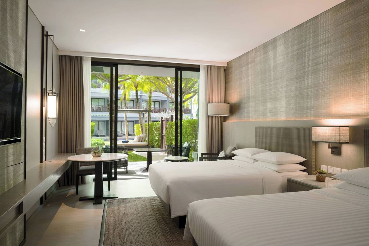 A modern hotel room with two beds, a dining area, and a balcony with a view of palm trees and buildings.