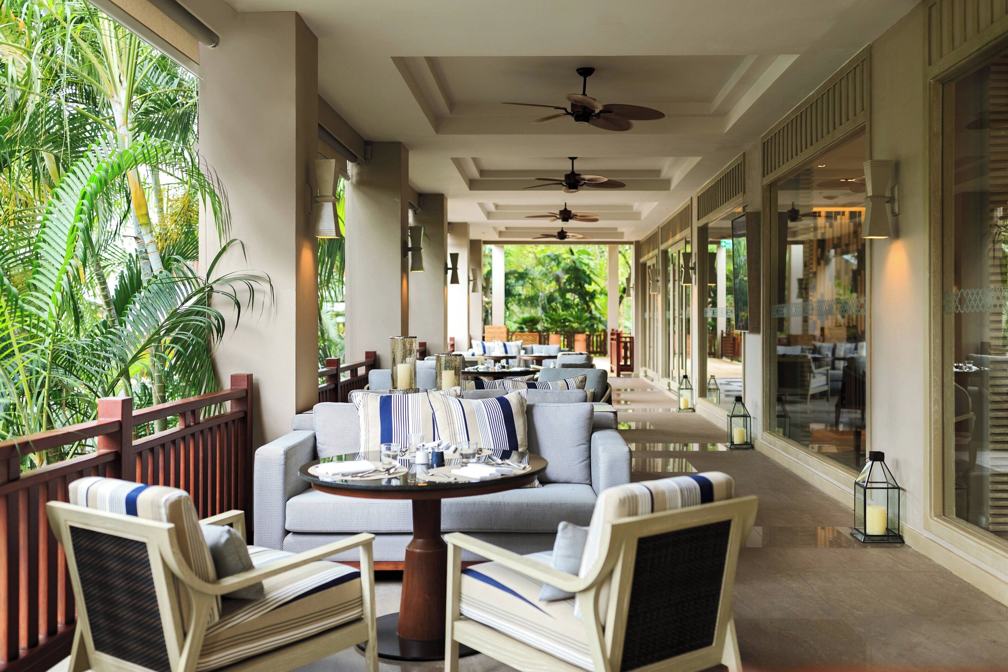 A spacious indoor area with a long table and several chairs, surrounded by large windows and a view of greenery.