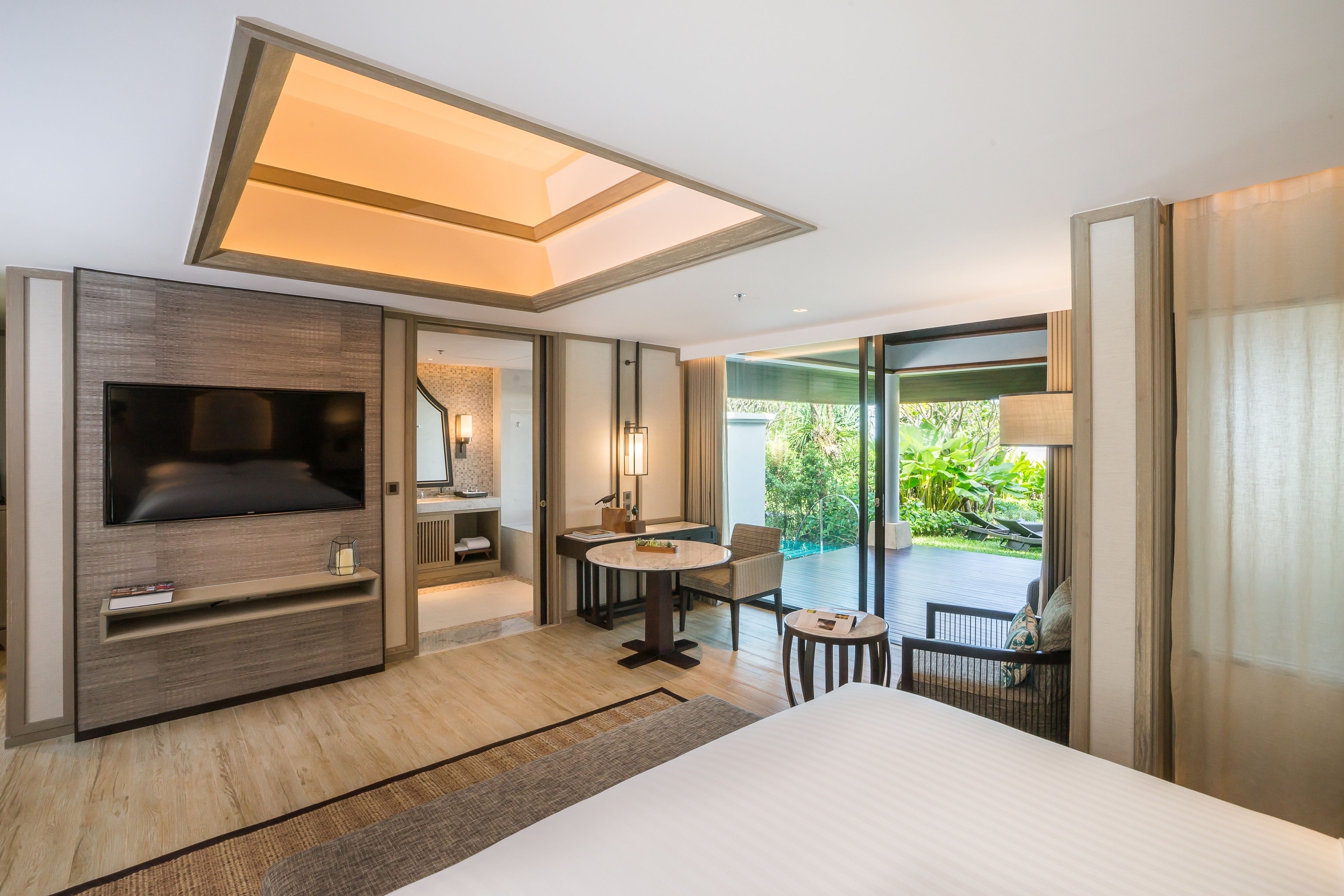A modern hotel room with a large bed, a flat-screen TV, a dining area with a table and chairs, and a balcony with a view of greenery.