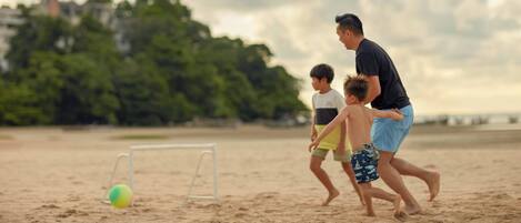 A sandy beach with a goalpost and a ball, and a person and two children playing near it.