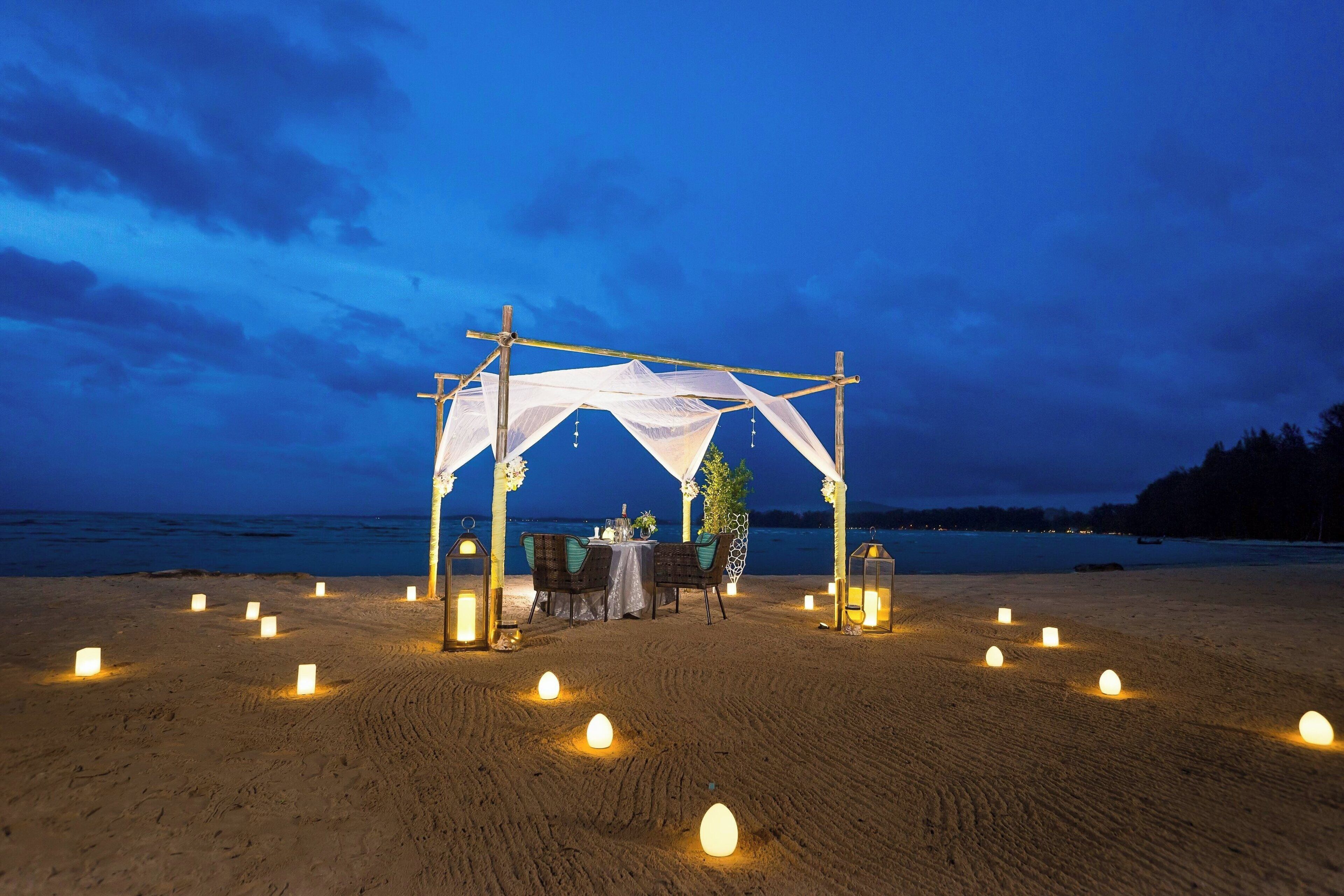 A beachside outdoor dining setup with a canopy, tables, chairs, and candles.