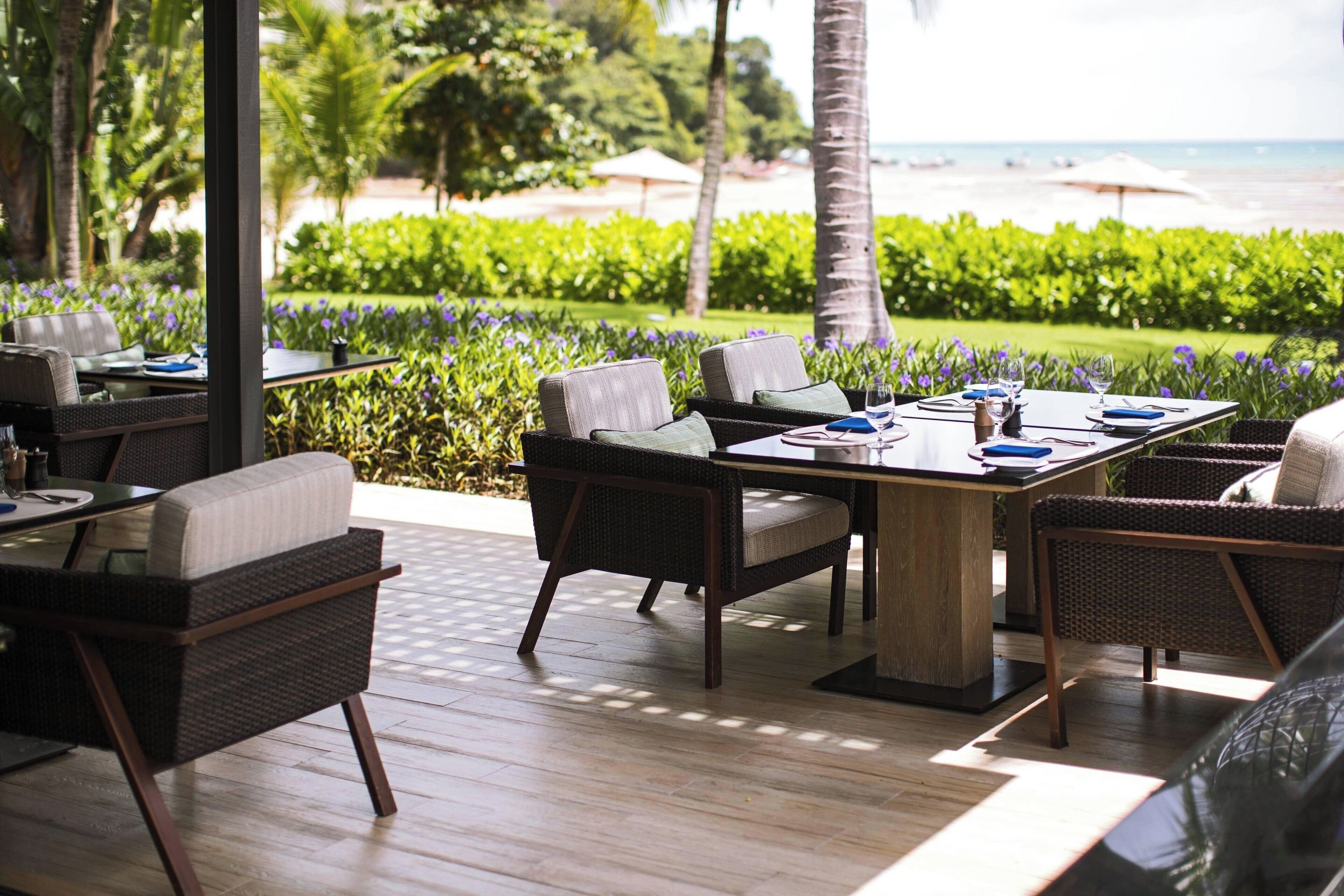 An outdoor dining area with wicker furniture, a wooden table, and a view of the beach and ocean.