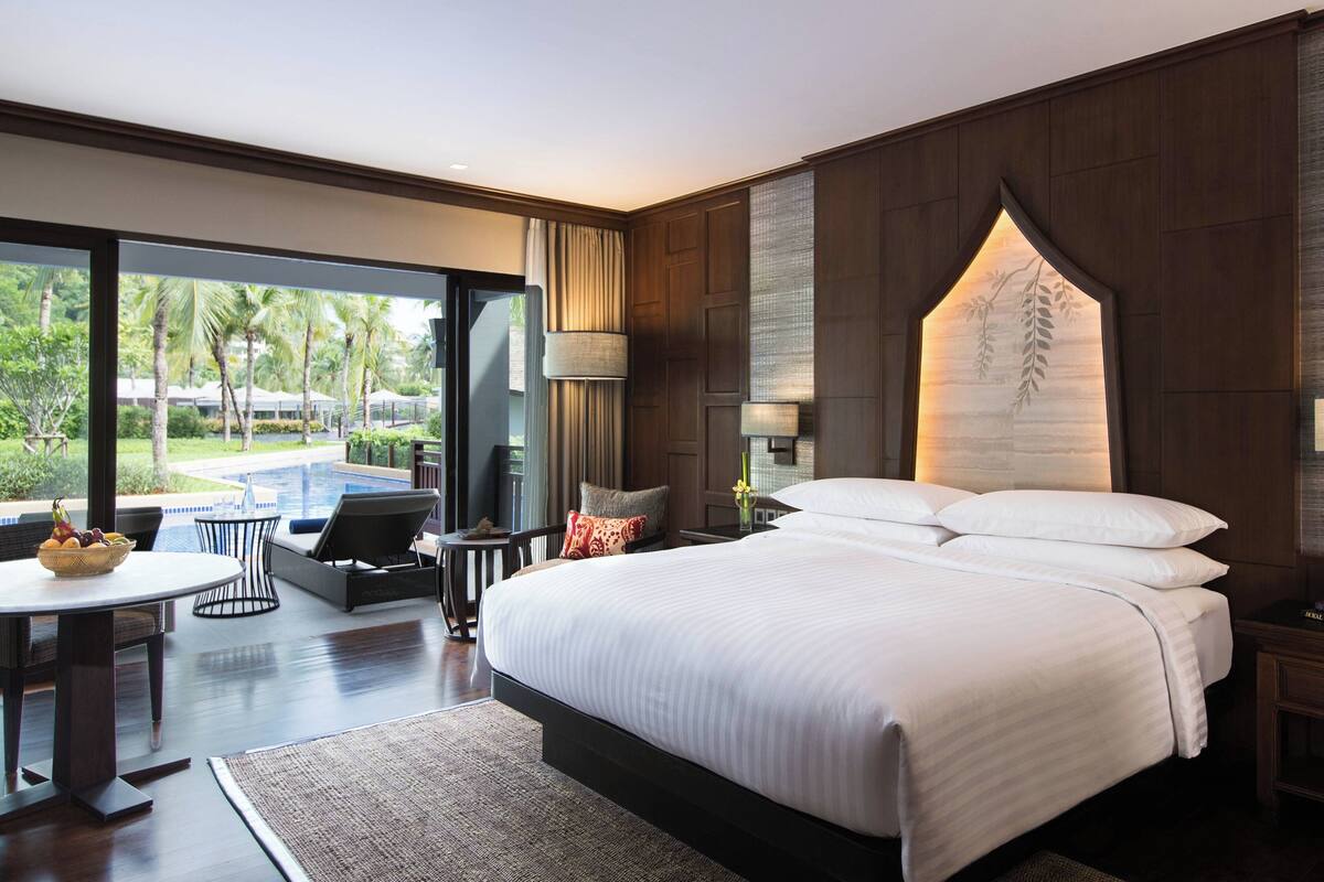 A modern hotel room with a large bed, a dining area with a table and chairs, and a view of a pool and greenery outside.
