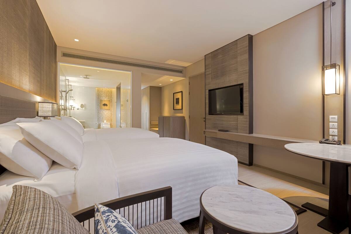 A modern hotel room with a large bed, a TV mounted on the wall, a round dining table, and a small seating area.