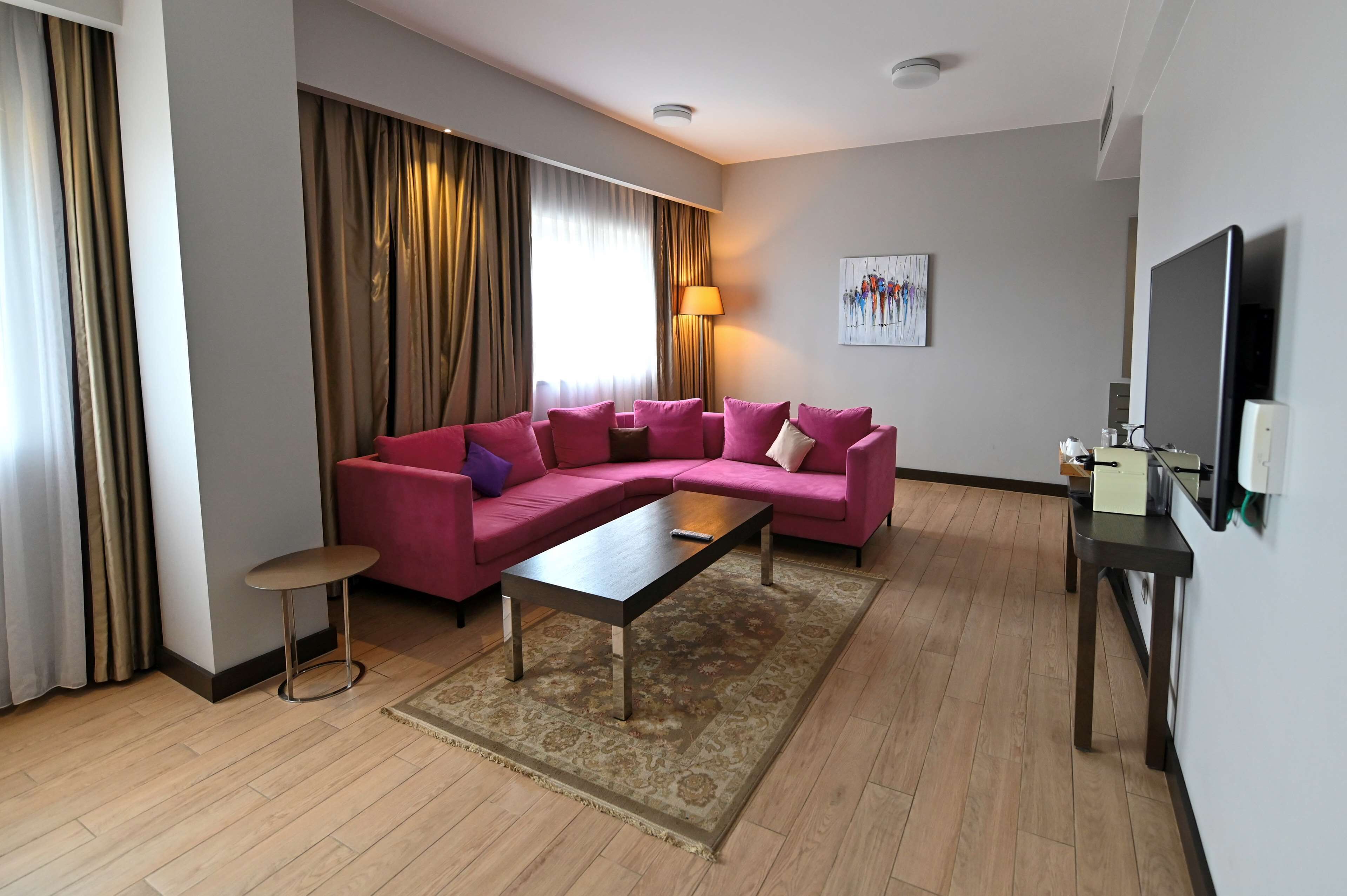 Ein modernes Hotelzimmer mit einem rosafarbenen Ecksofa, einem kleinen runden Tisch, einem Flachbildfernseher und einem Gemälde an der Wand.