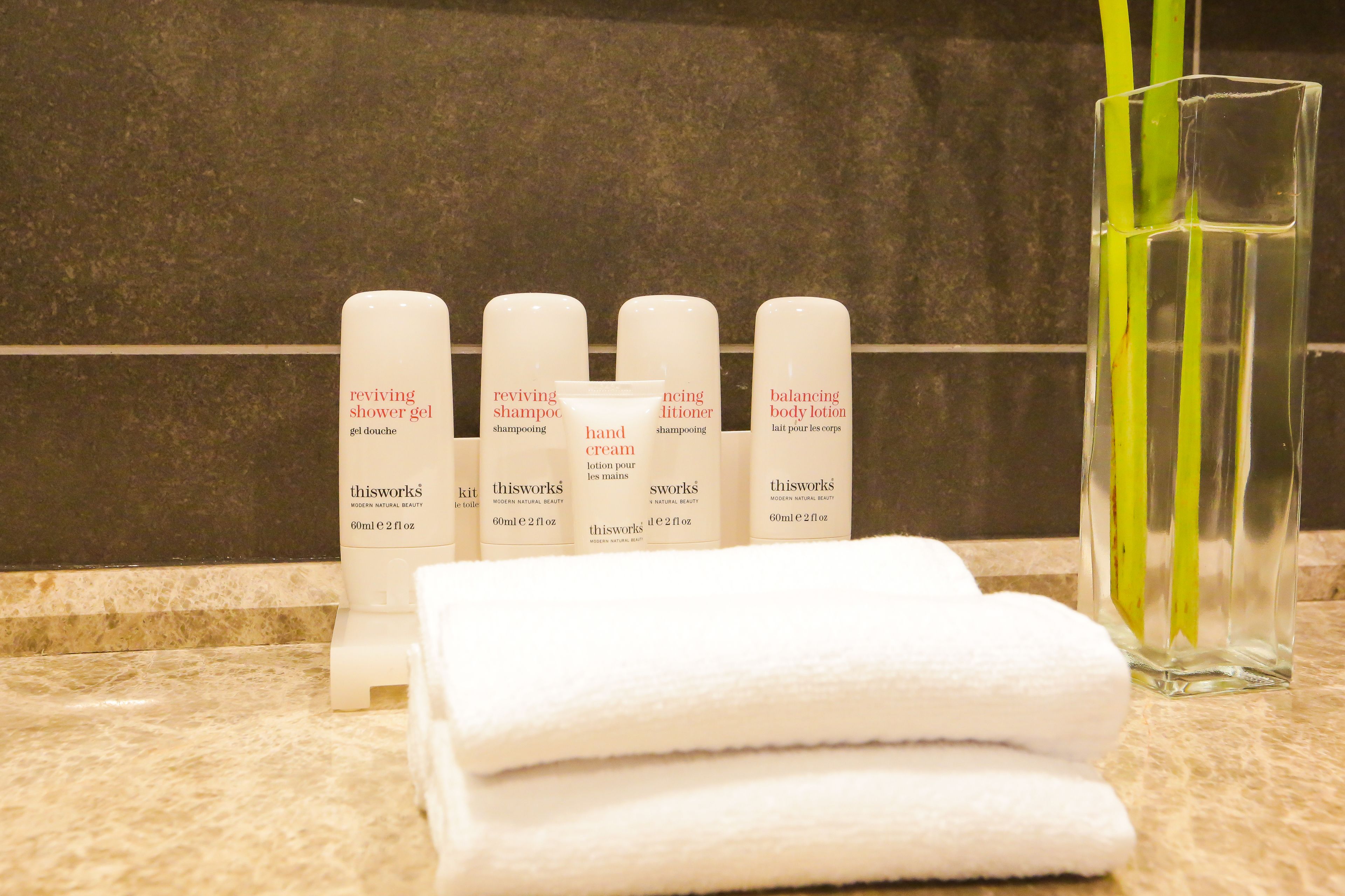 A bathroom counter with toiletries including a shower gel, shampoo, hand cream, and body lotion, along with a glass of water and a towel.