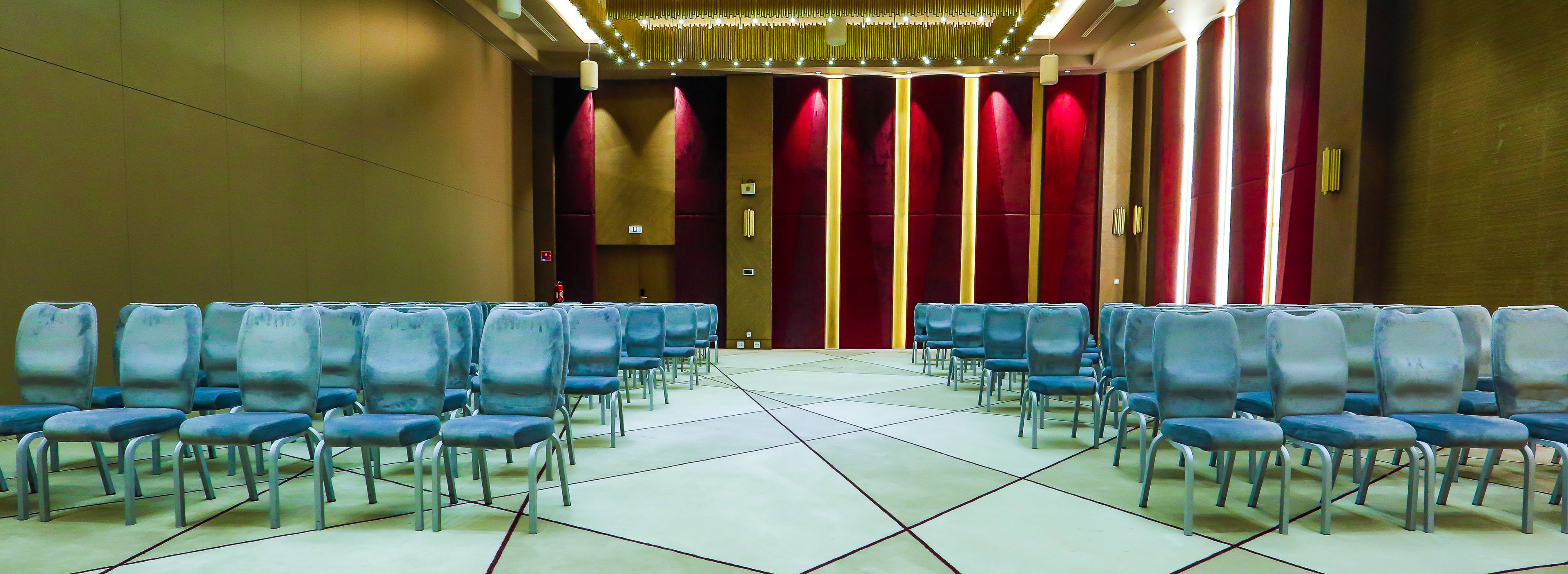 A conference room with rows of blue chairs, a tiled floor, and a red curtain backdrop.
