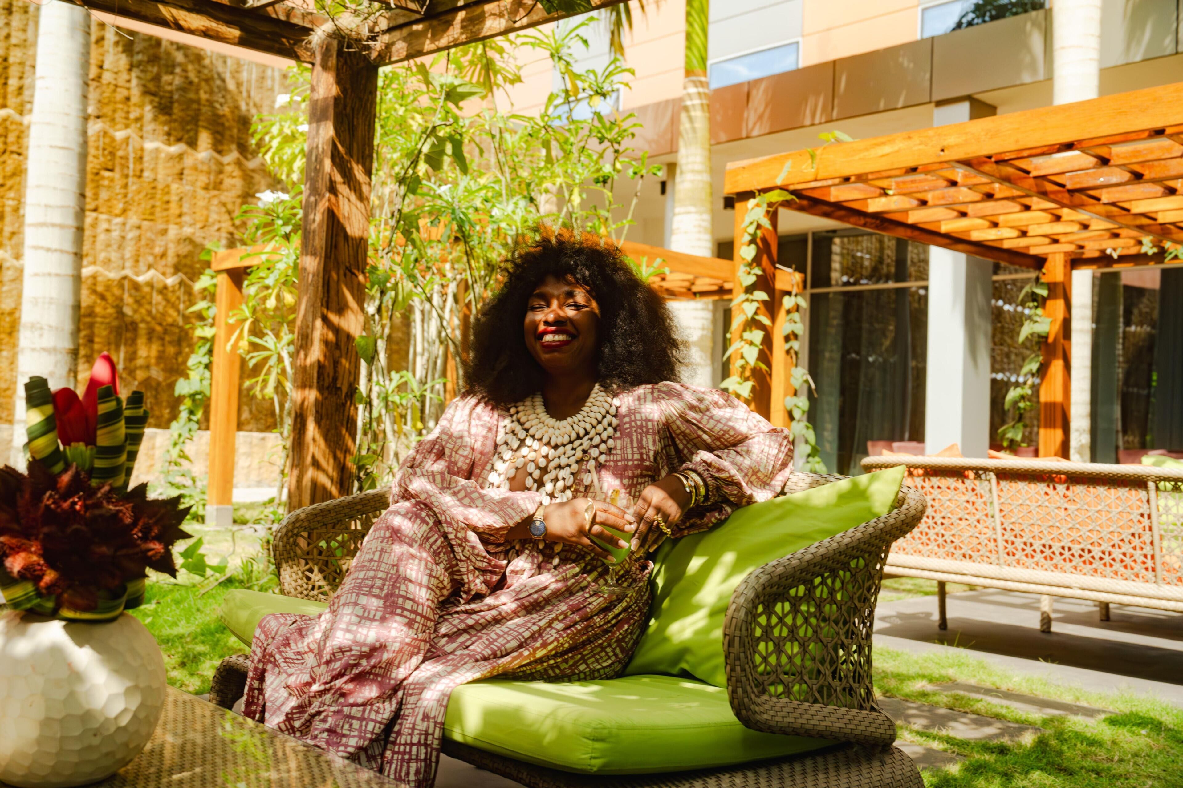 A person seated on a wicker chair with a green cushion, surrounded by lush greenery and a wooden pergola.