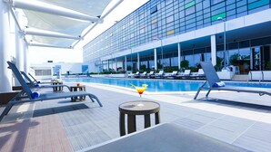 Un espace piscine moderne avec des chaises longues, un bar et un grand bâtiment en verre en arrière-plan.