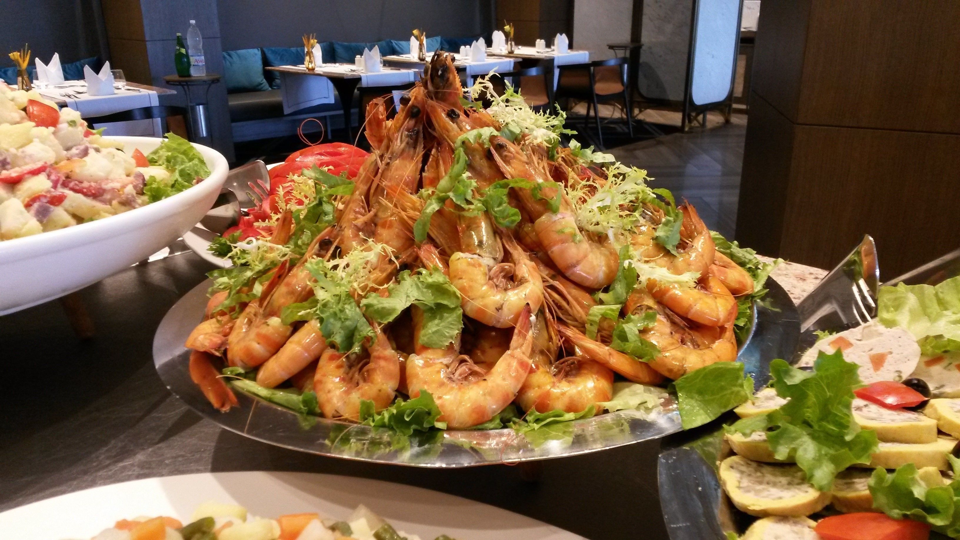 Ett buffébord med ett stort fat fullt av kokta räkor, garnerat med grönsaker, omgivet av andra rätter, inklusive sallader och skivad frukt.