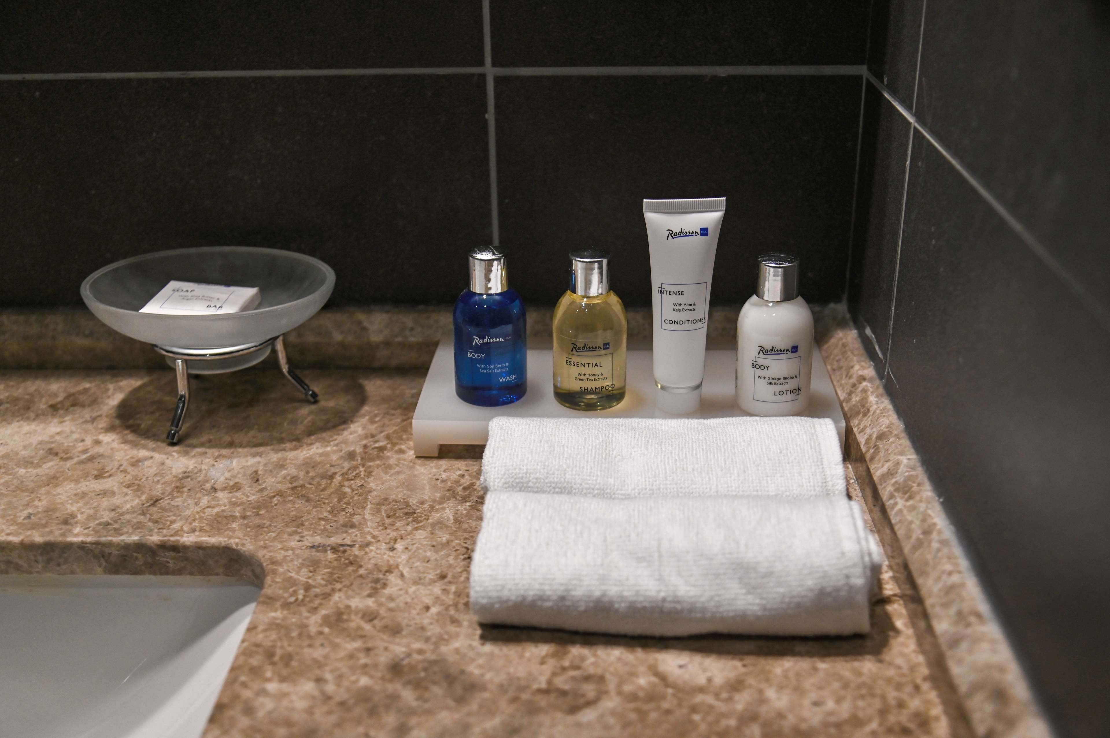 Una encimera de baño con un lavabo, tres botellas de gel de baño, champú y loción Radisson, y una toalla doblada.