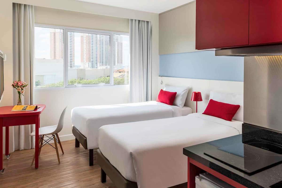 A hotel room with two beds, a desk, a chair, a red cabinet, and a view of buildings through the window.