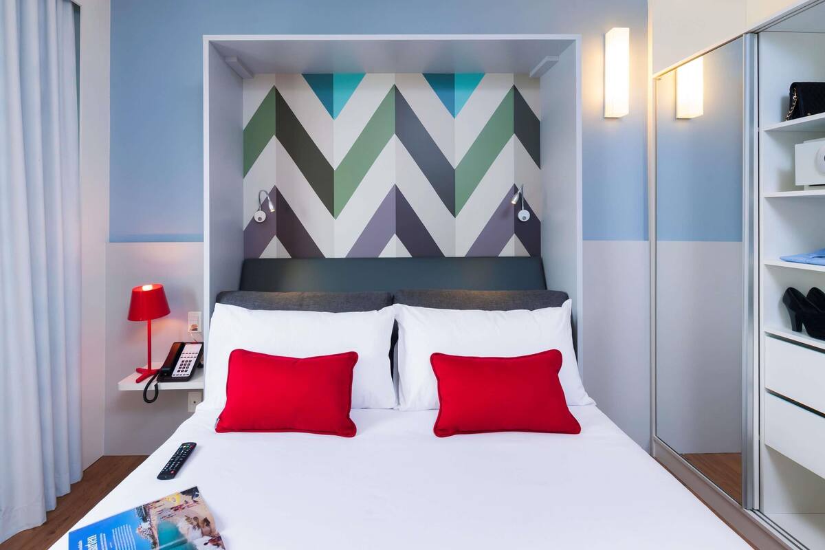 A hotel room with a bed, red pillows, a phone on a bedside table, and a wall with a geometric pattern.