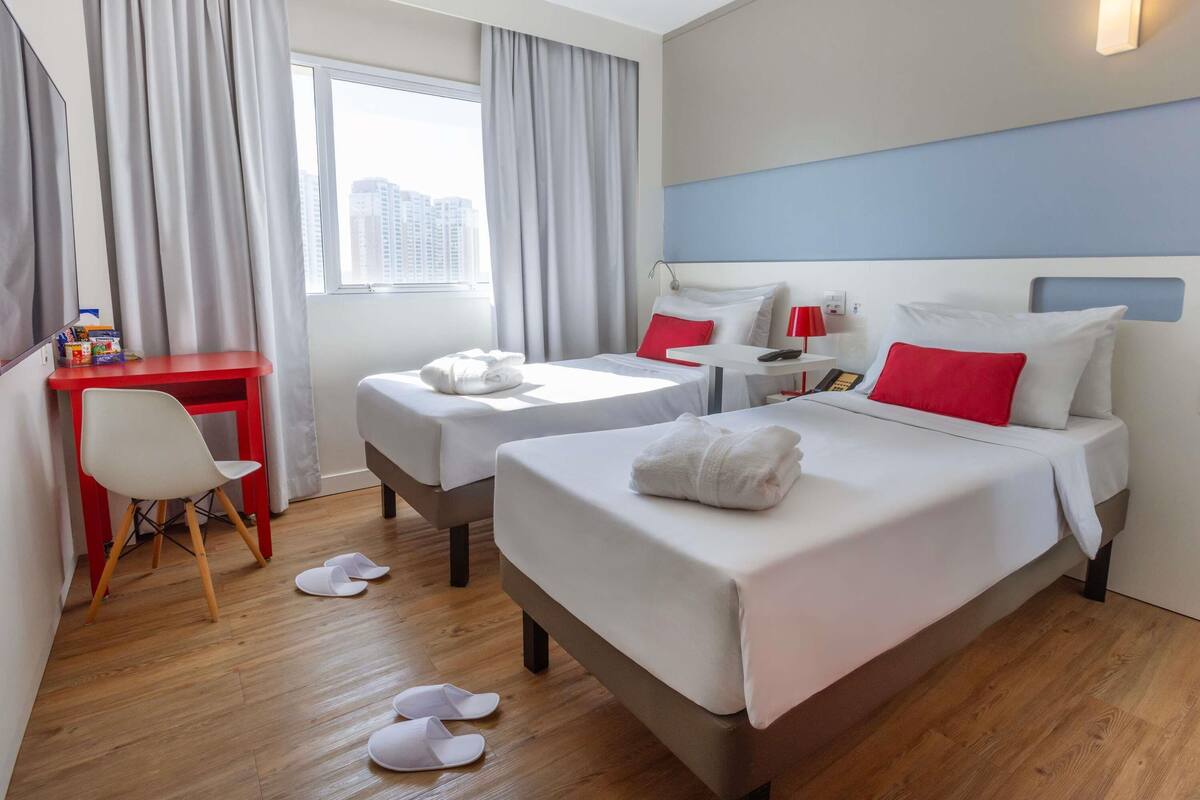 A hotel room with two beds, a desk, a chair, and a view of the city.
