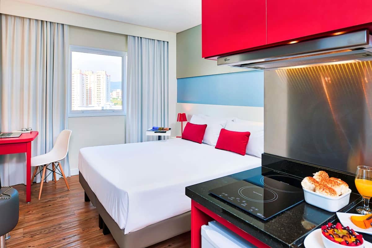 A hotel room with a bed, a desk, a chair, a window with curtains, a kitchenette with a stove, and a breakfast tray.