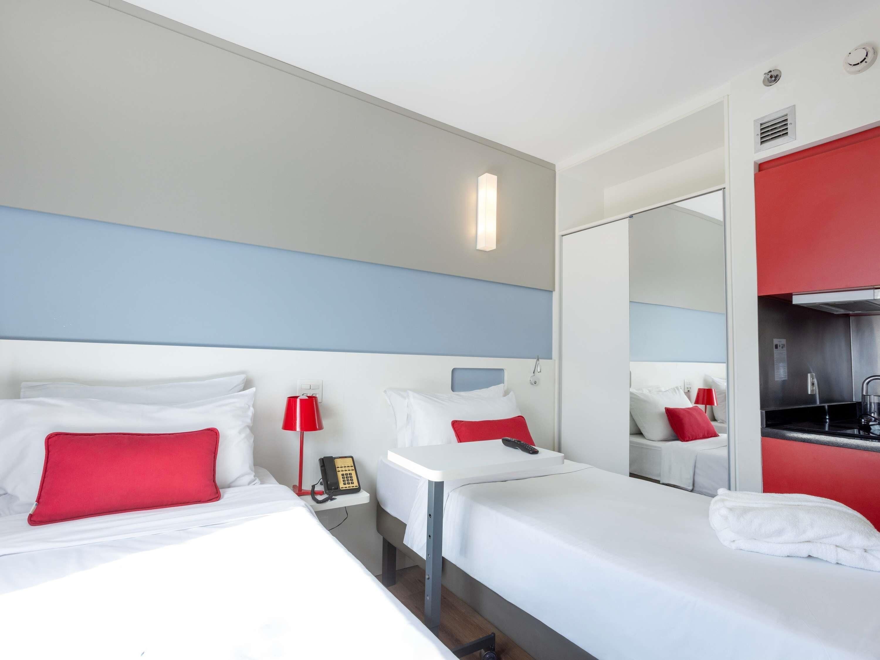 A hotel room with two beds, a red lamp, a telephone, and a mirror.