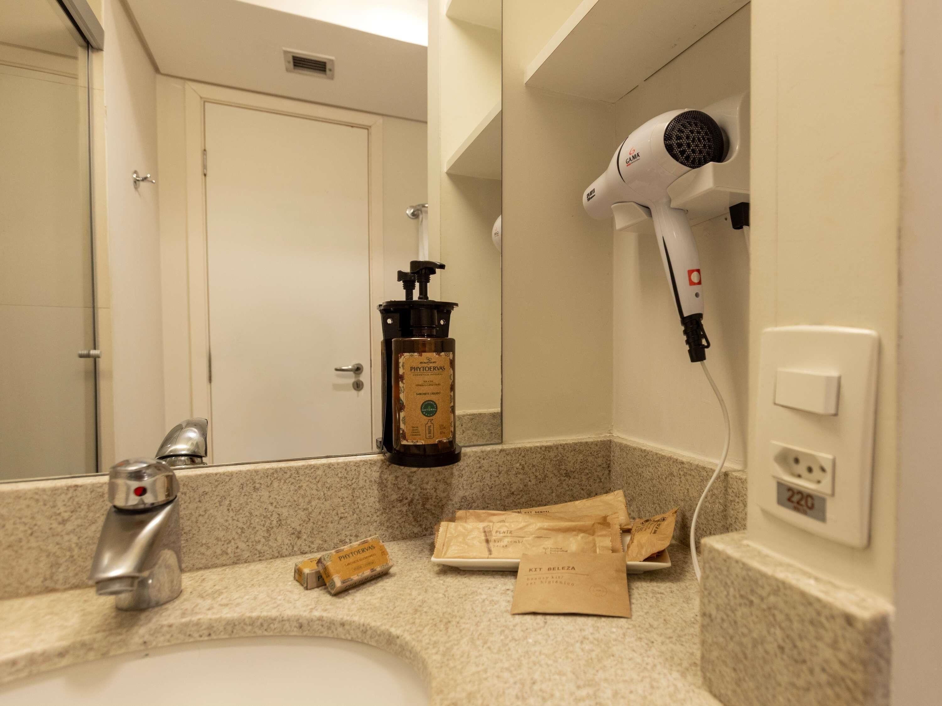 A bathroom with a sink, a hairdryer, a bottle of hand soap, and a towel dispenser.