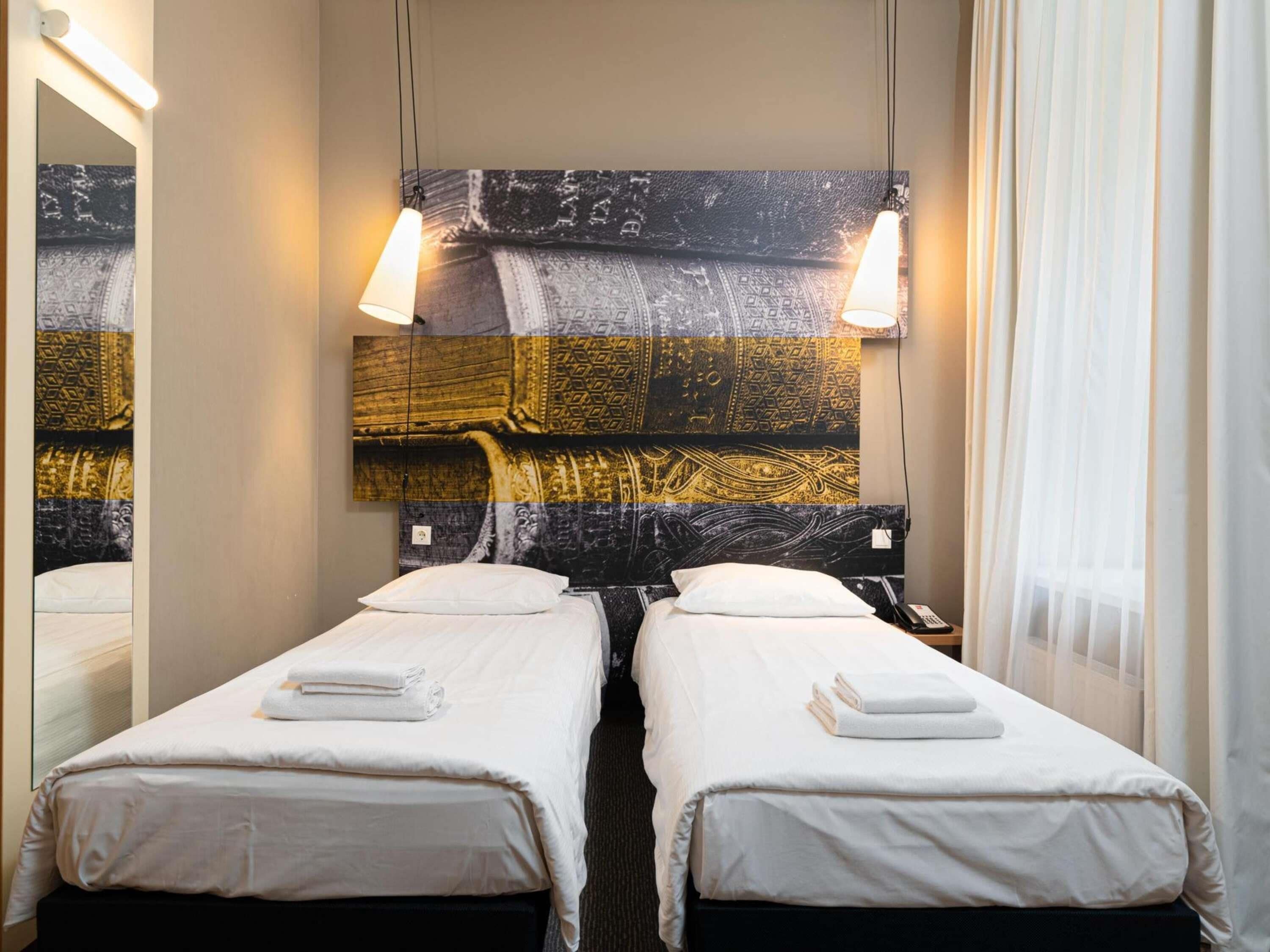 A hotel room with two single beds, a large wall tapestry, and a hanging light fixture.